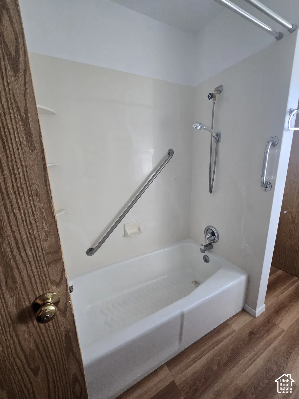 Bathtub with shower fixture, grab bars, wood-finish flooring, and light-colored wall surround