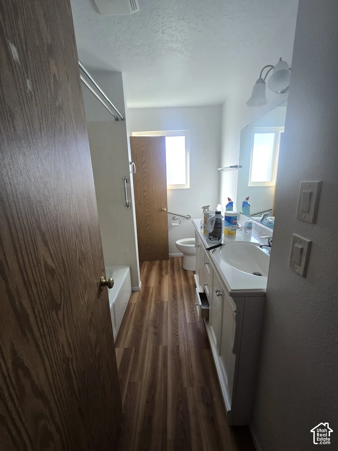 Wood-finish flooring, white vanity with integrated sink, wall-mounted light fixture, and white bathtub with shower rod