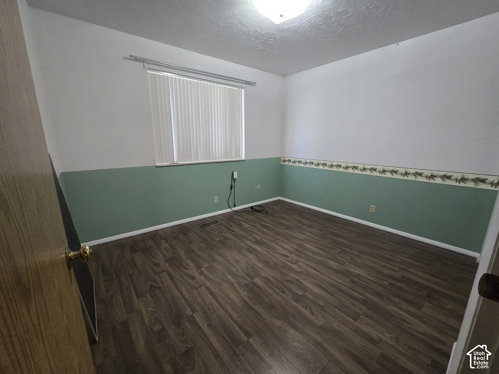Wood-finish flooring with white baseboards, wall-mounted light fixture, and vertical blinds on the window