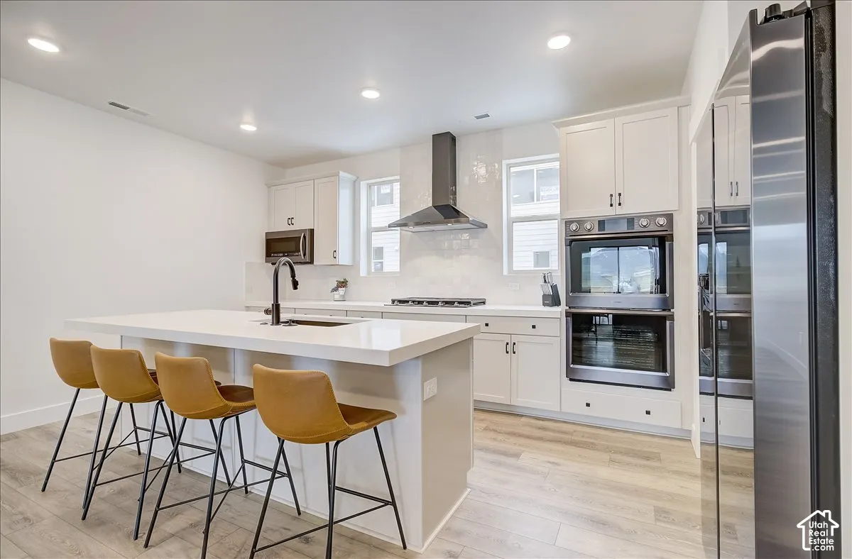Kitchen featuring a large center island, white cabinetry, integrated stainless steel appliances, a black range hood, and light wood-finish flooring