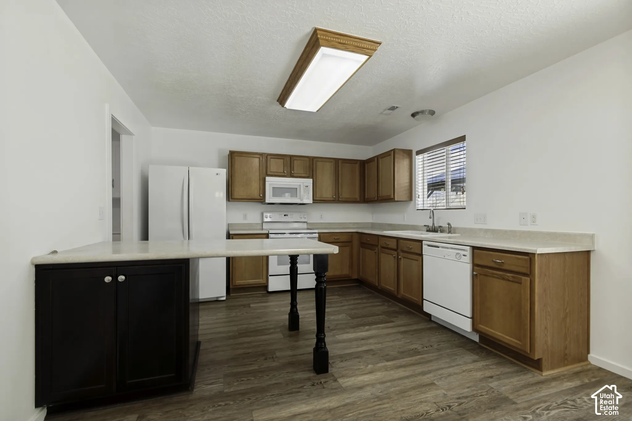 Kitchen featuring wood-finish flooring, light wood cabinetry, light countertops, white appliances, and a breakfast bar with dark wood base