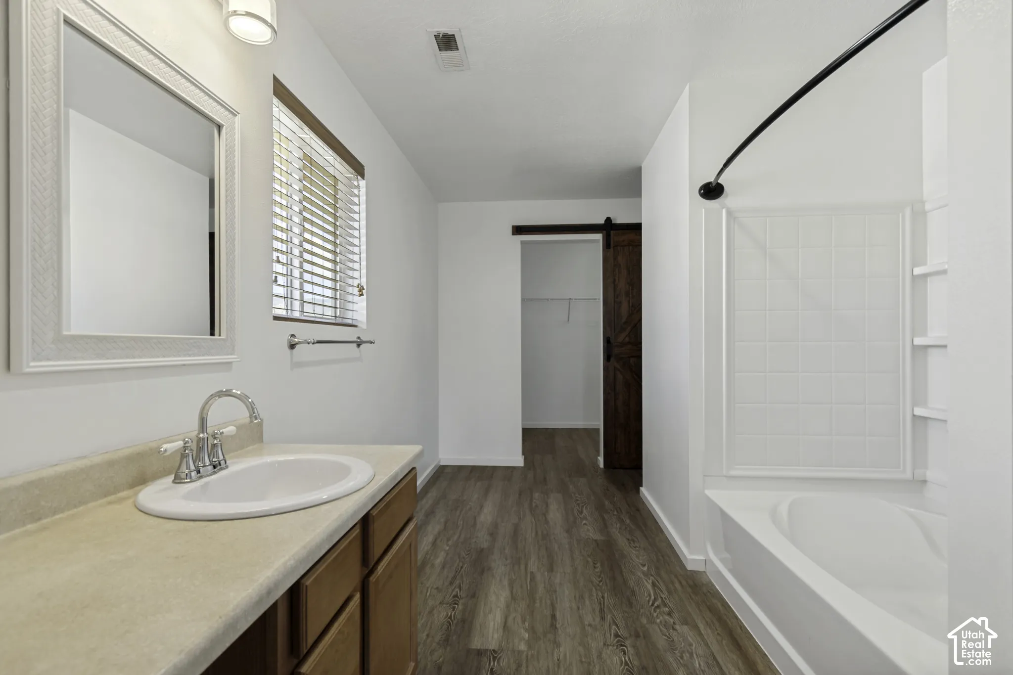 White vanity with built-in sink, wood-finish flooring, white bathtub with tiled surround, and sliding barn door