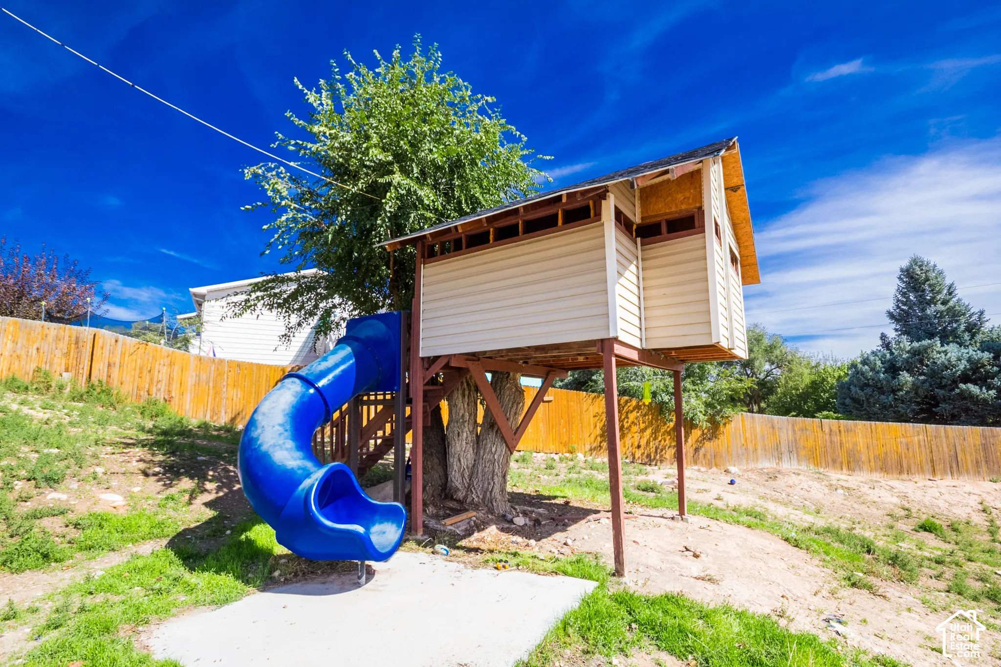 Outdoor play structure with a blue slide, light-colored siding, and a sloped grassy area