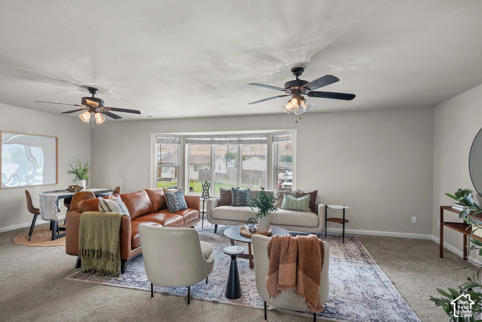 Bay window with multiple panes, two ceiling fans with integrated lighting, neutral-toned wall paint, and carpet flooring
