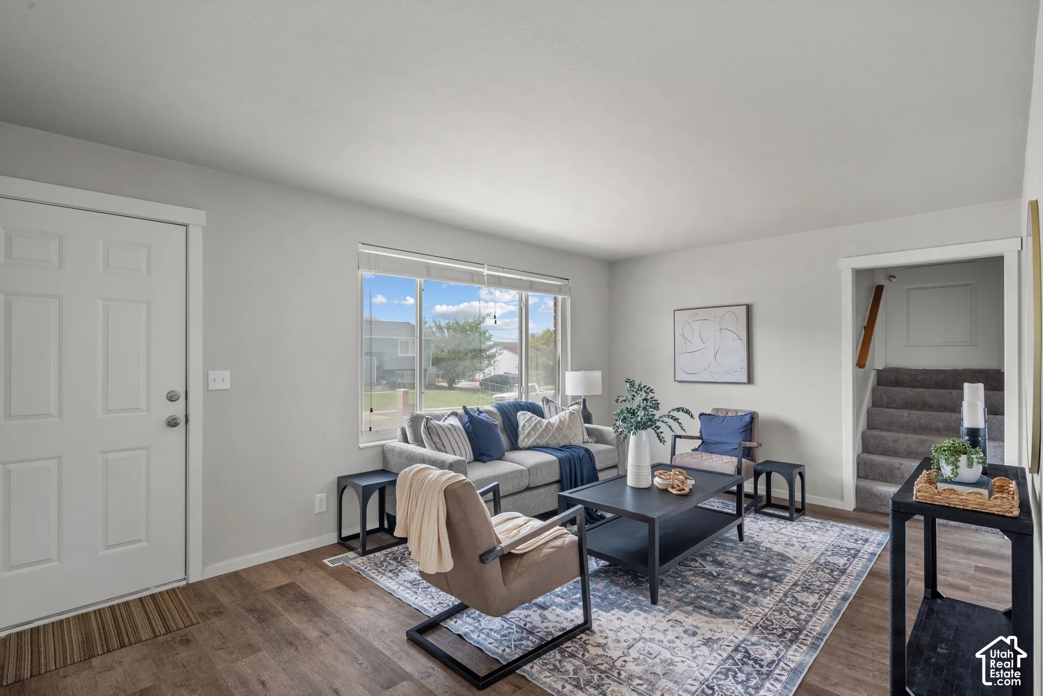 Spacious living area featuring wood-finish flooring, a large window, light-toned walls, and a carpeted staircase
