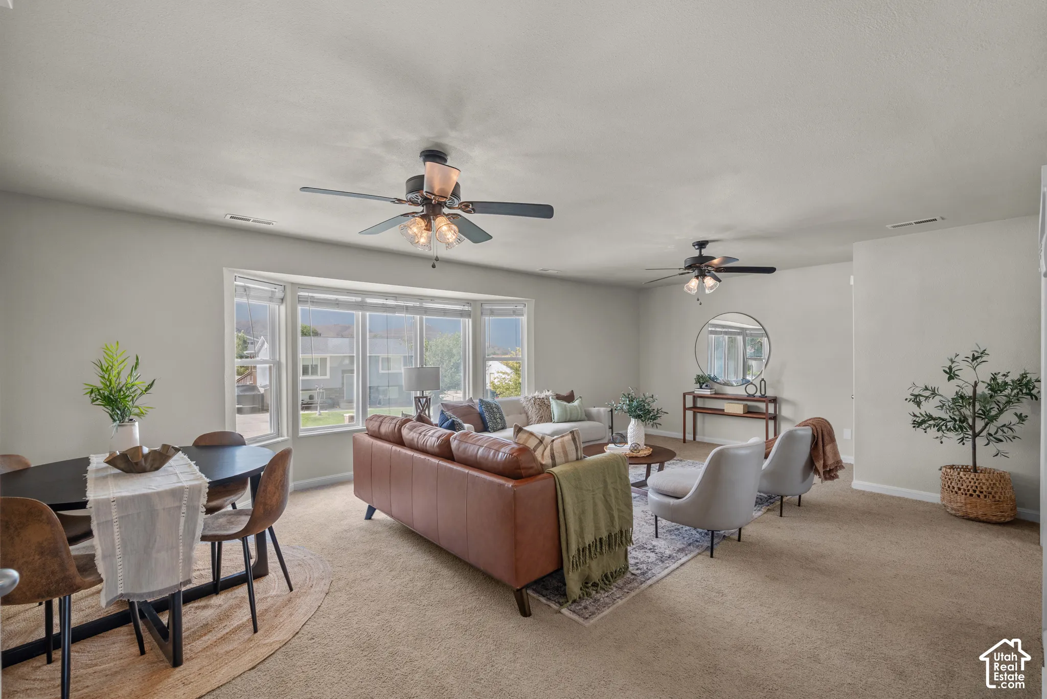 Open-concept living area with bay windows, two ceiling fans with integrated lighting, and neutral carpeting throughout