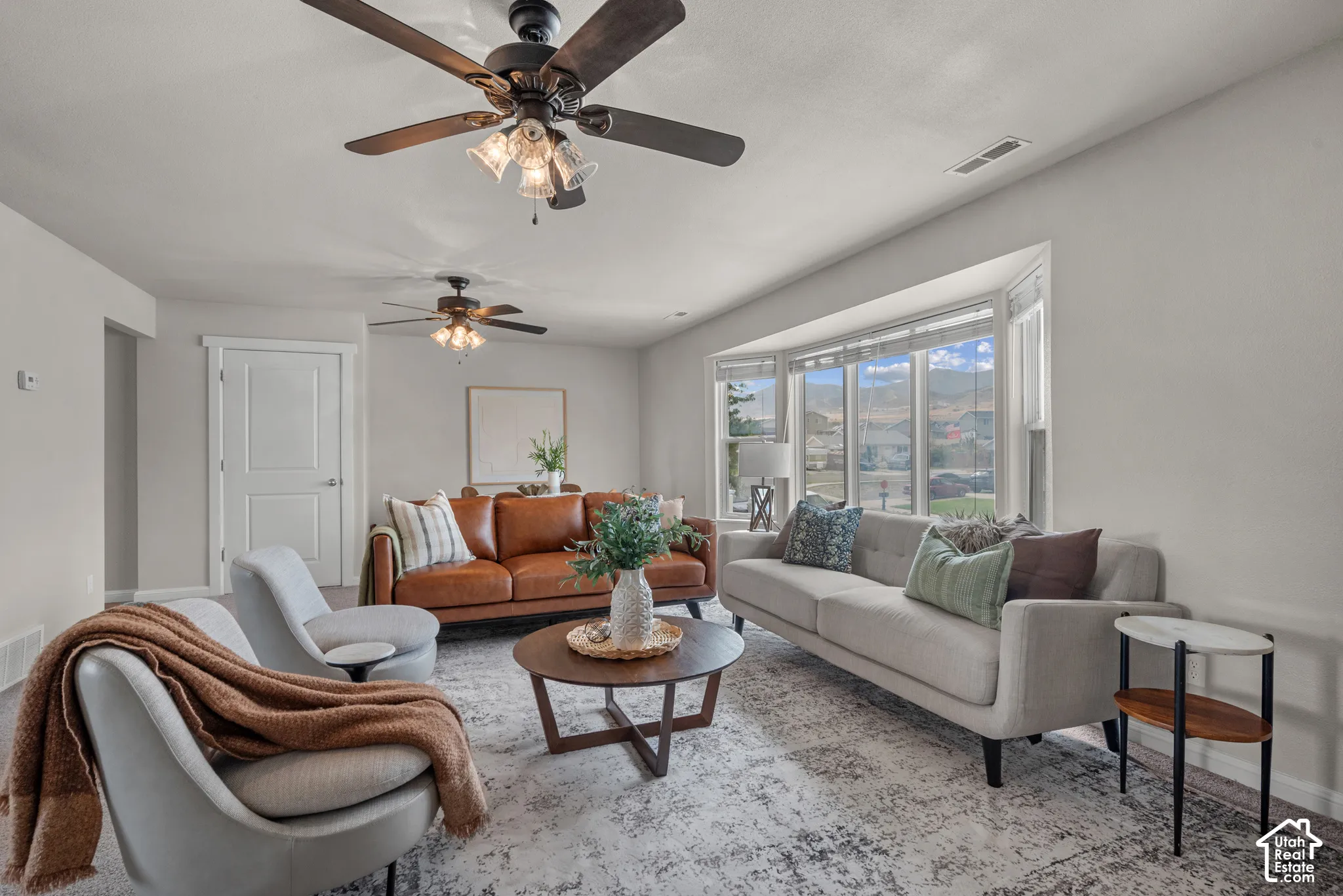 Spacious living area featuring a large bay window, light-toned walls, and multiple ceiling fans
