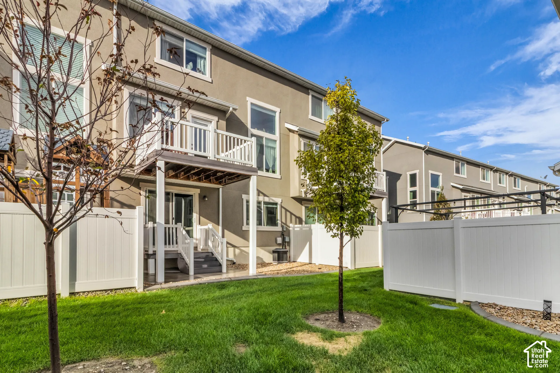 Manicured lawn with a young tree, white privacy fencing, and a ground-level patio with stairs leading to a second-story balcony