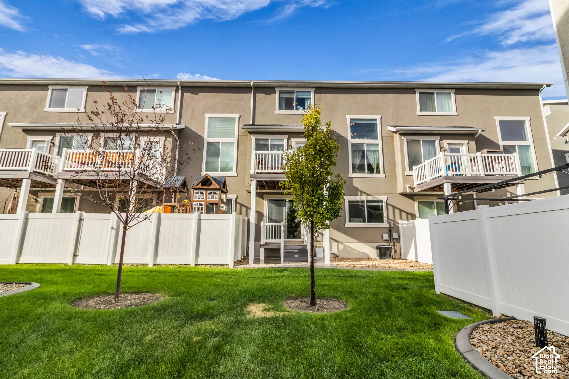 Stucco exterior with multiple balconies, featuring white privacy fencing, manicured lawn, and mature trees