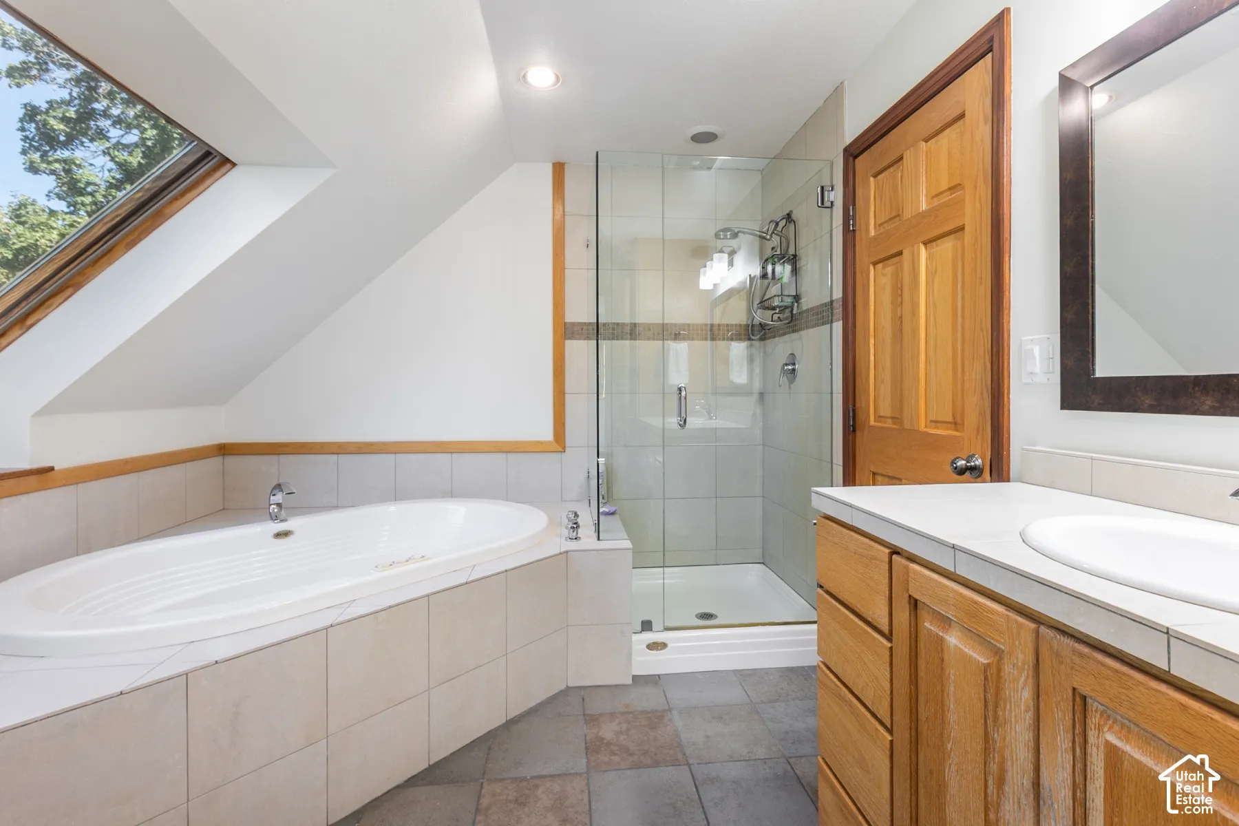 Soaking tub with tile surround, glass-enclosed shower, wood-finish vanity with countertop, and skylight