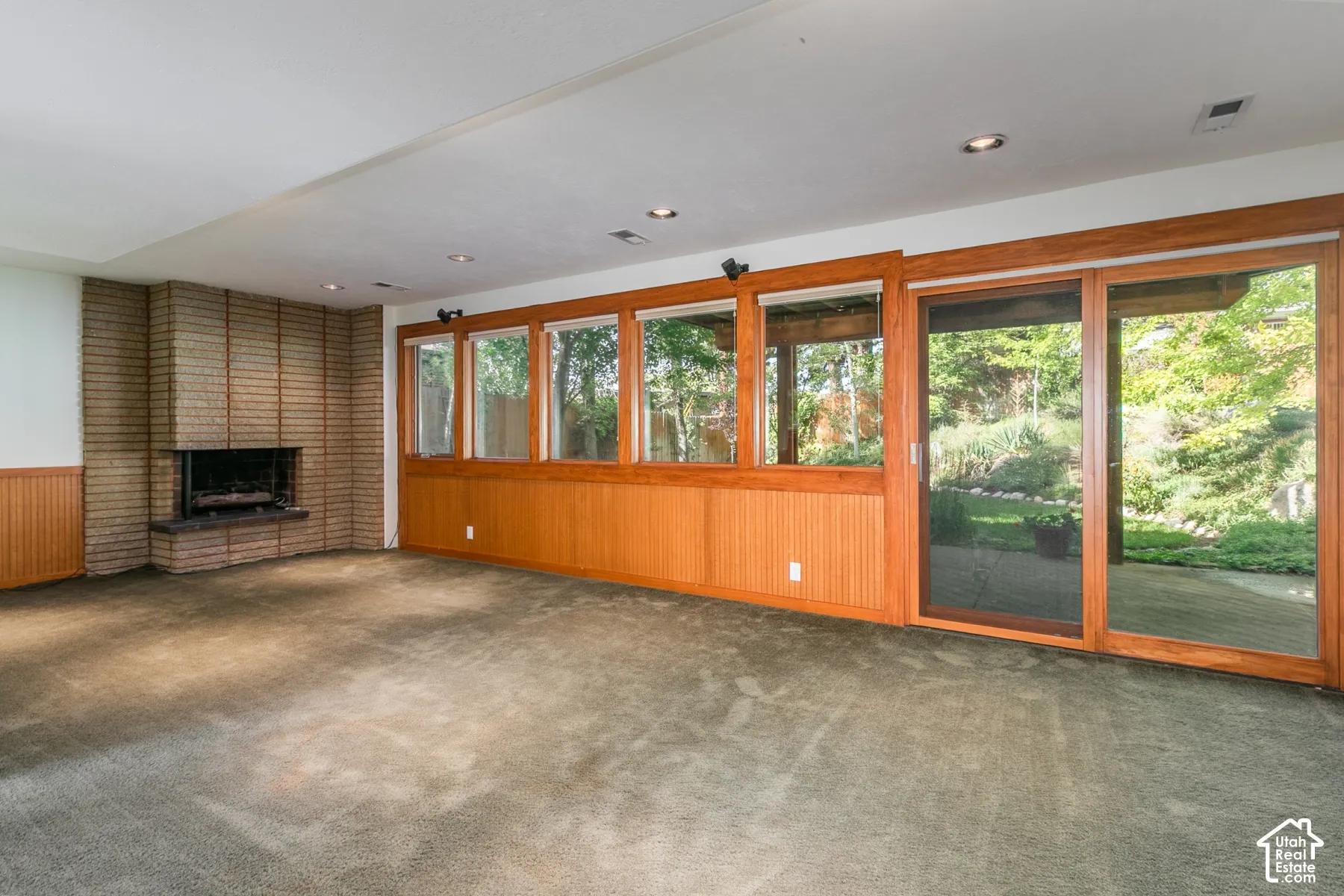 Brick fireplace with hearth, wood-paneled wainscoting, recessed lighting, expansive glass doors, and multiple windows