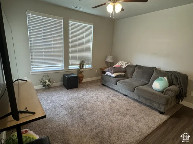 Carpeted living area featuring two windows with blinds, a ceiling fan with integrated lighting, and wood-finish flooring