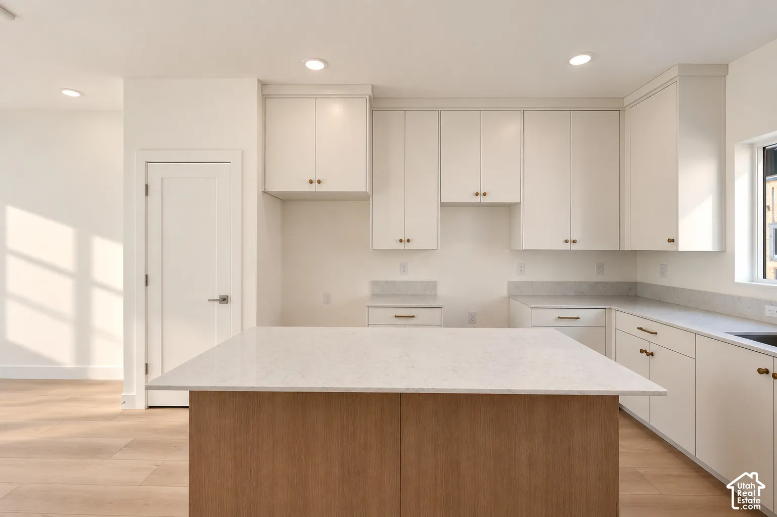 Central kitchen island with a light-toned countertop, white cabinetry with brass-finish hardware, light-toned wood-finish flooring, recessed lighting, and a single window