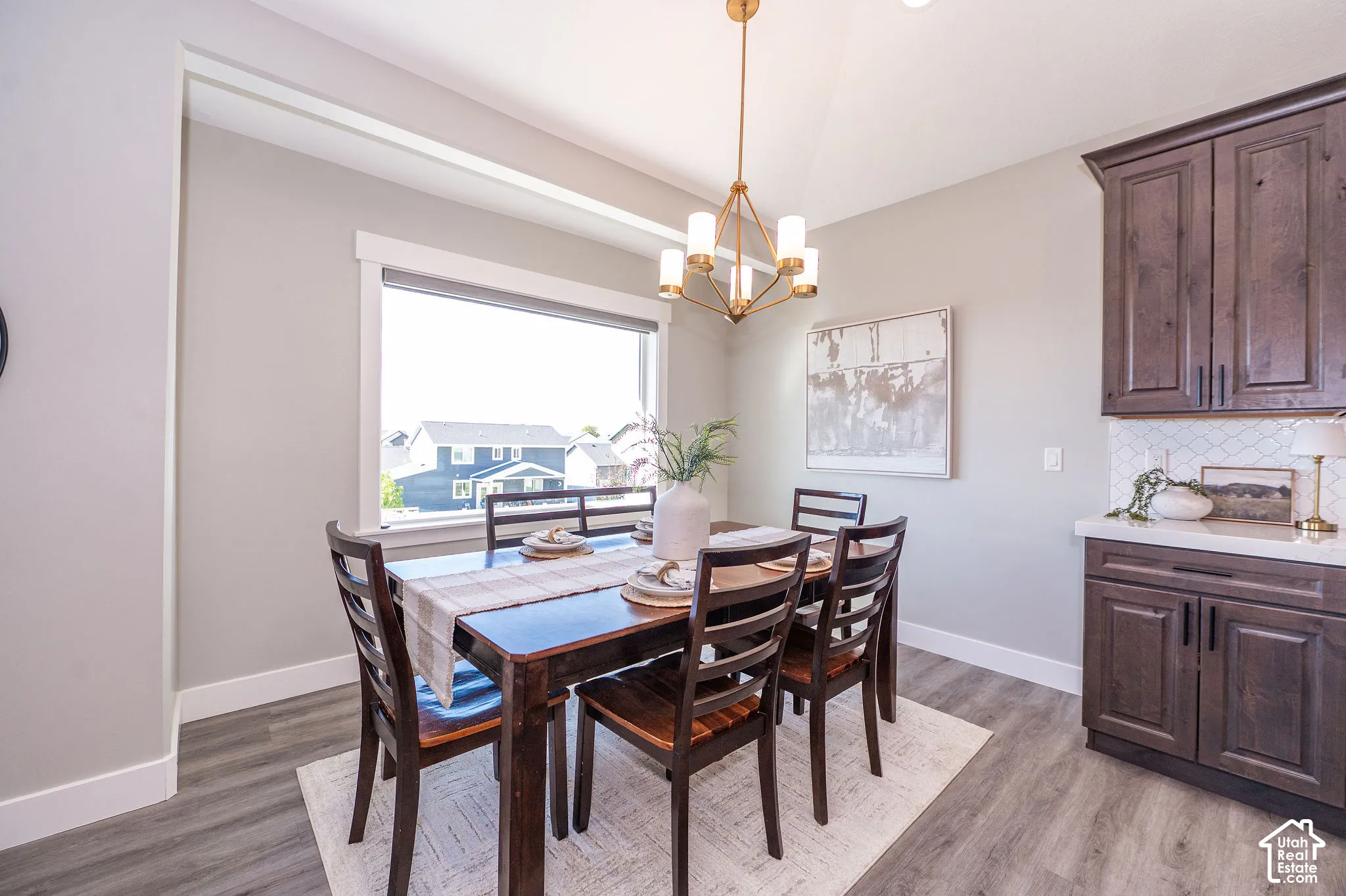 Dining area with wood-finish flooring, a large window, a modern chandelier, and dark wood cabinetry with white countertops and a tiled backsplash