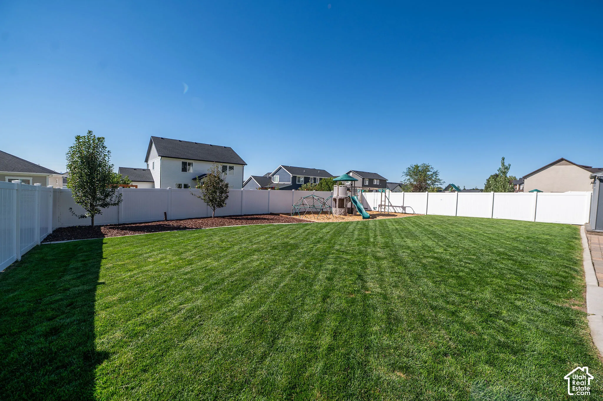 Expansive backyard featuring a manicured lawn, established trees, perimeter fencing, and a paved patio area