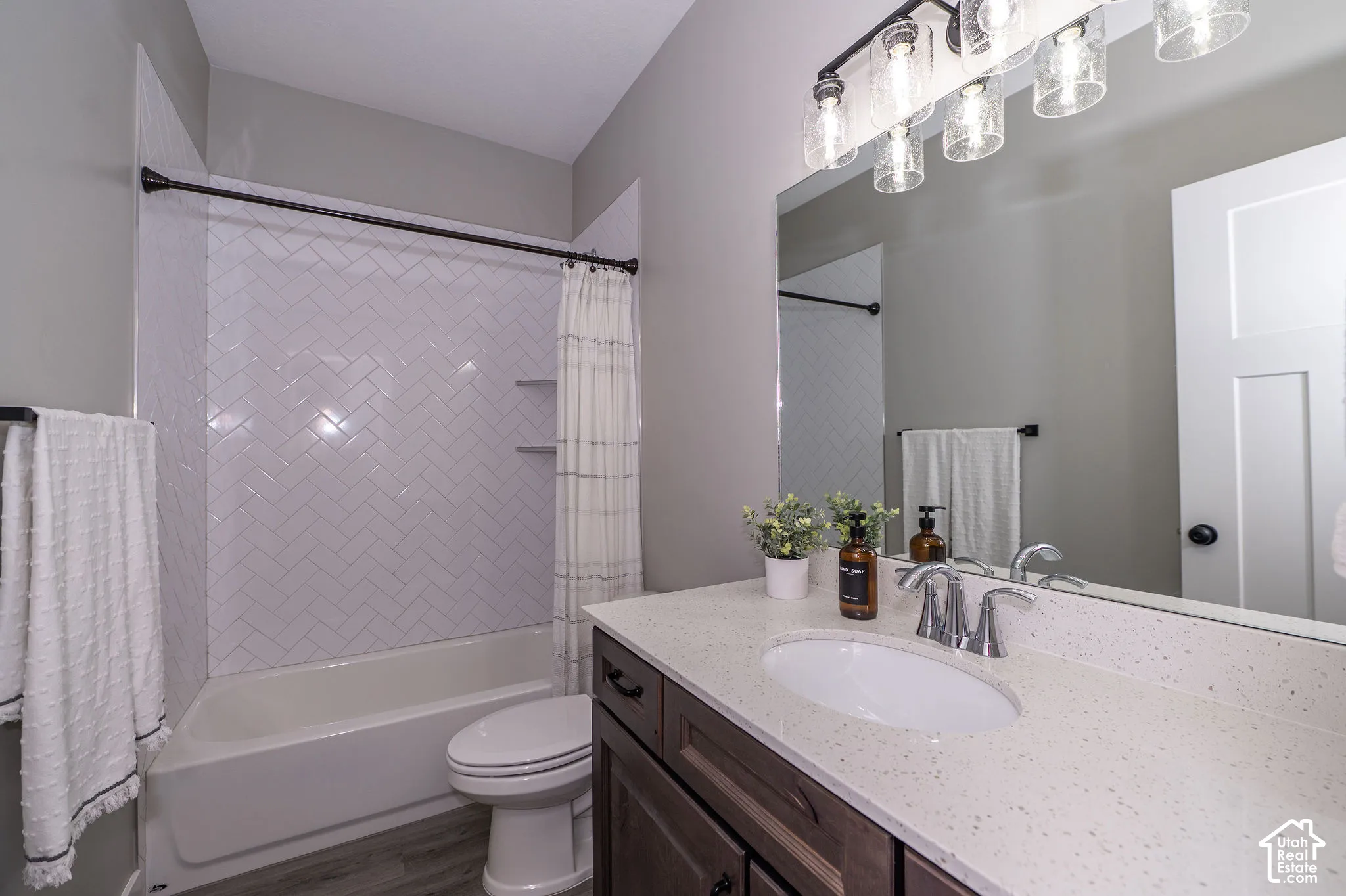 White subway tile with herringbone pattern, white bathtub, wood-finish vanity with built-in sink, light speckled countertop, and wall-mounted vanity lighting