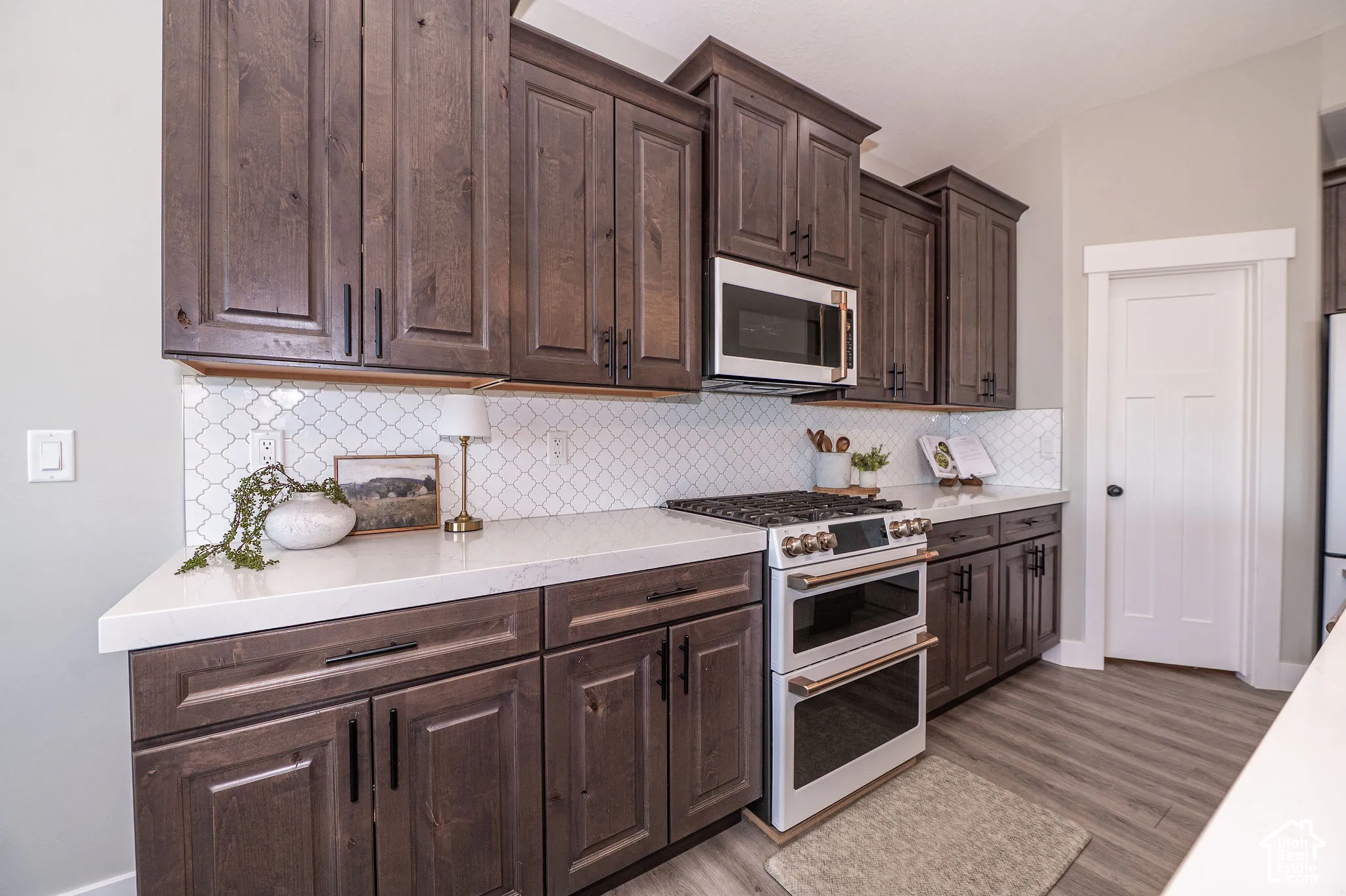 Dark wood-finish cabinetry with modern hardware, white solid surface countertops, white arabesque tile backsplash, stainless steel microwave, and a double oven range