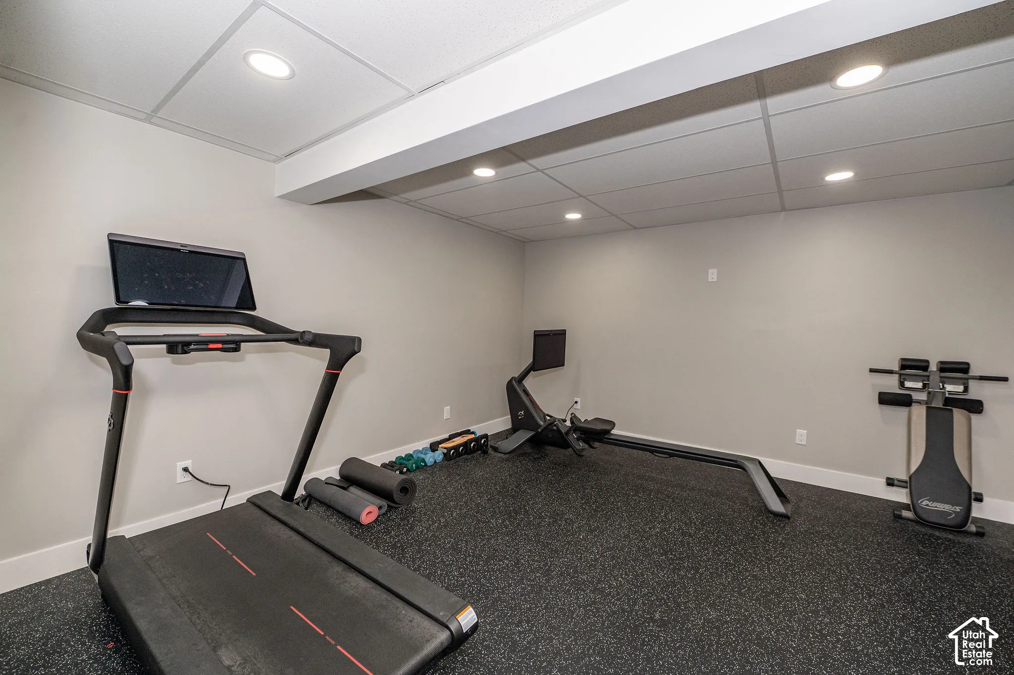 Recessed lighting illuminating a spacious room with a drop ceiling, featuring light-toned walls and durable rubber flooring