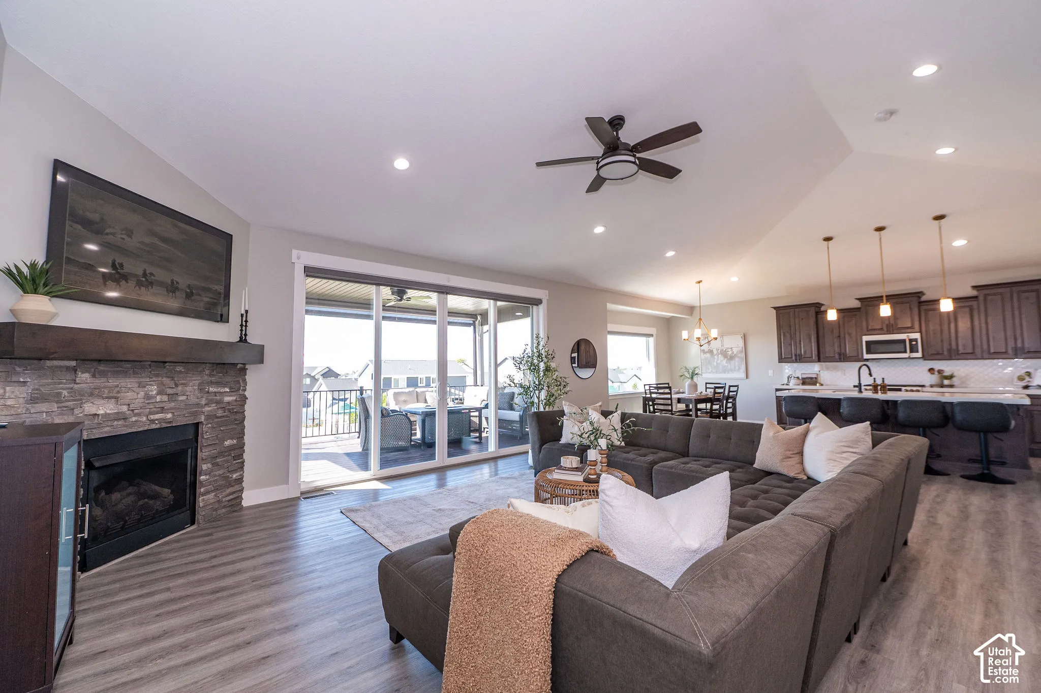 Vaulted ceiling, stone-surround fireplace with wood mantel, wood-finish flooring, sliding glass patio doors, and kitchen with dark wood cabinetry