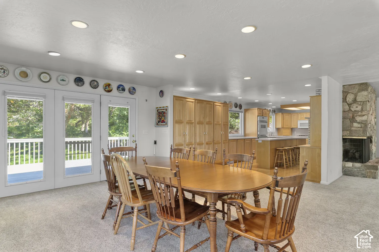Recessed lighting illuminating the room, multiple glass-paned doors opening to an outdoor deck, a natural stone fireplace, and light wood cabinetry in the kitchen area