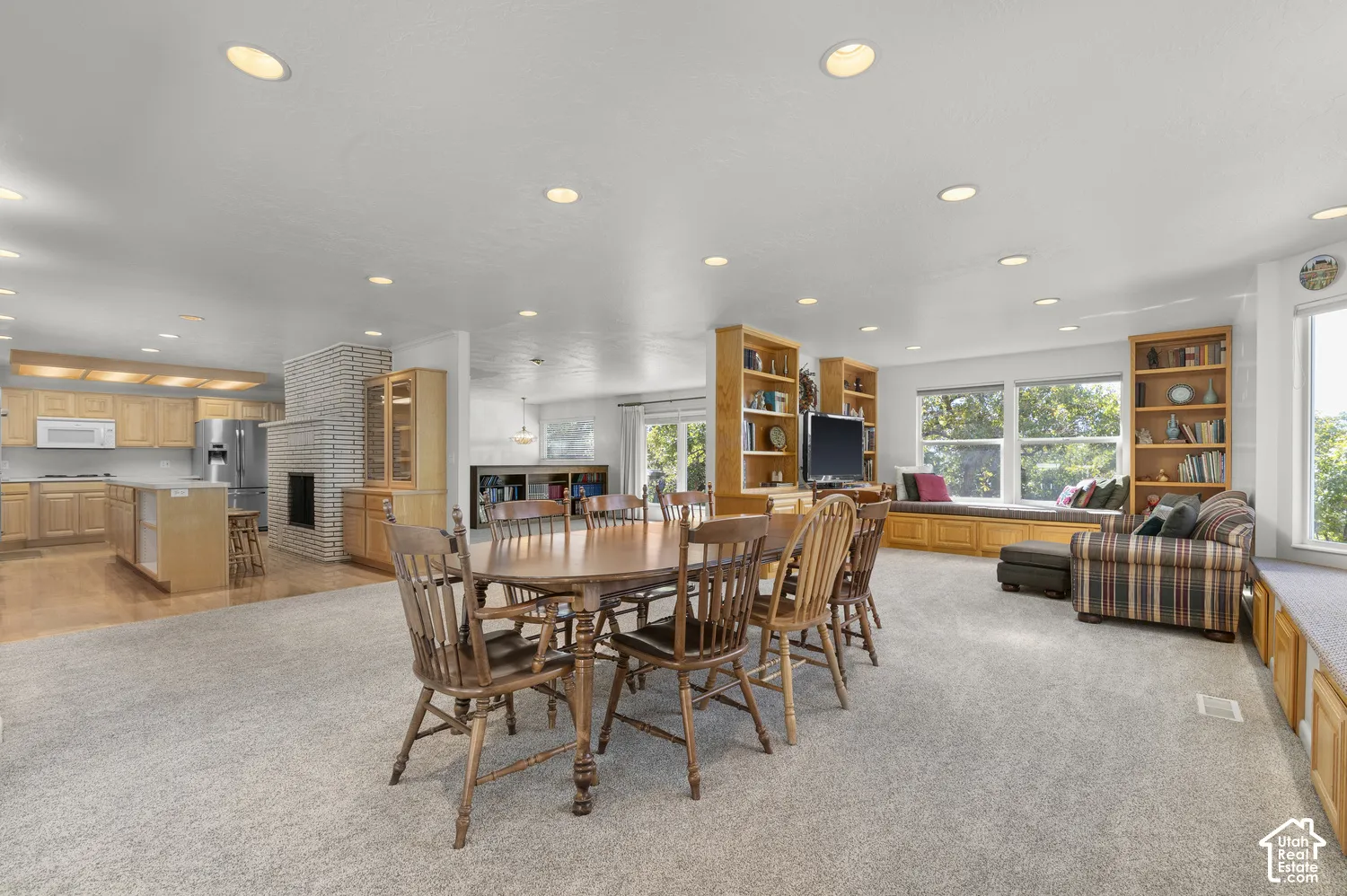 Recessed lighting throughout, built-in shelving, expansive windows, kitchen with light wood-finish cabinetry, and a brick fireplace