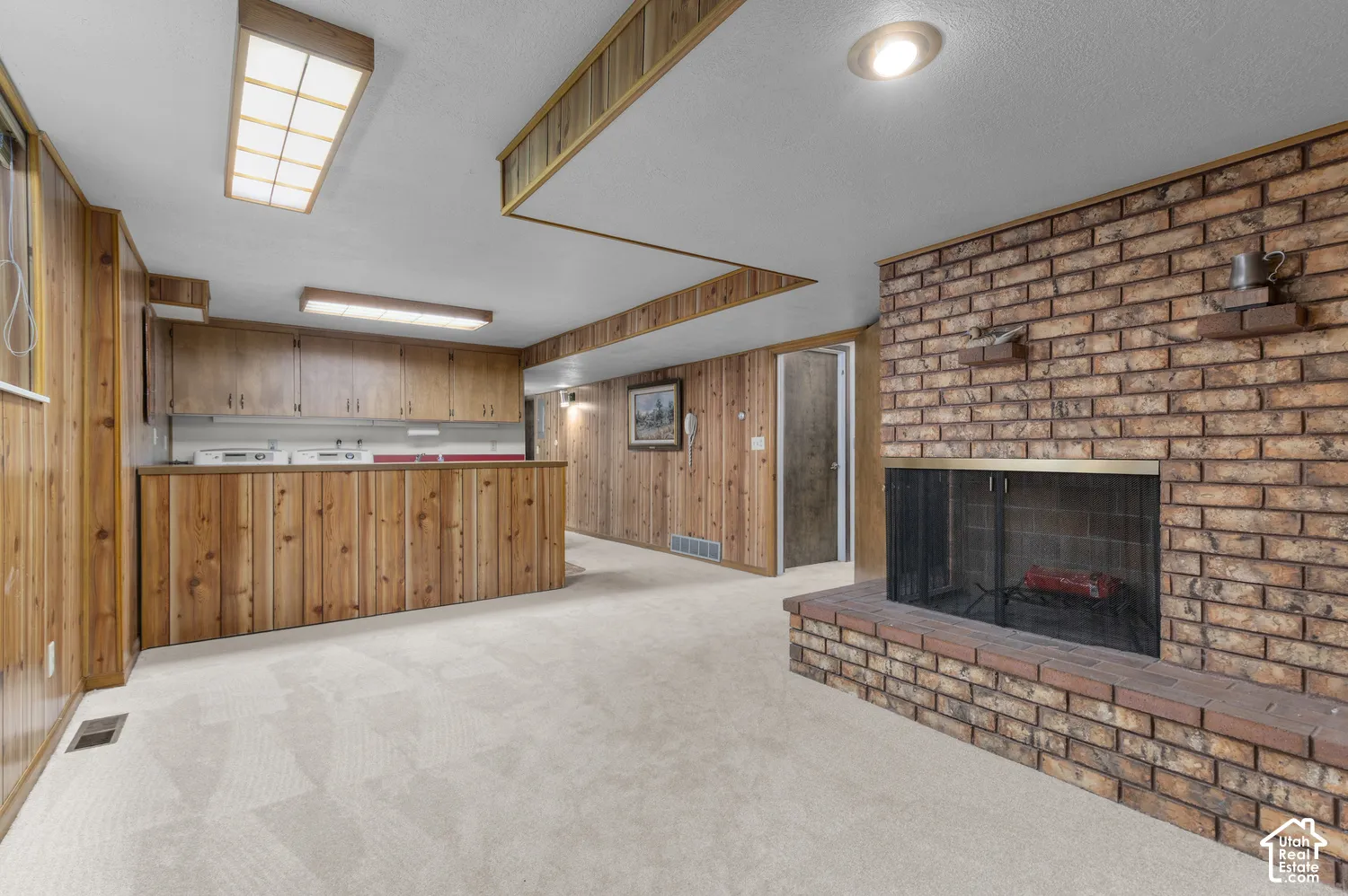 Brick fireplace with a hearth, light-toned carpet, wood paneling, wood-finish cabinetry, and recessed lighting