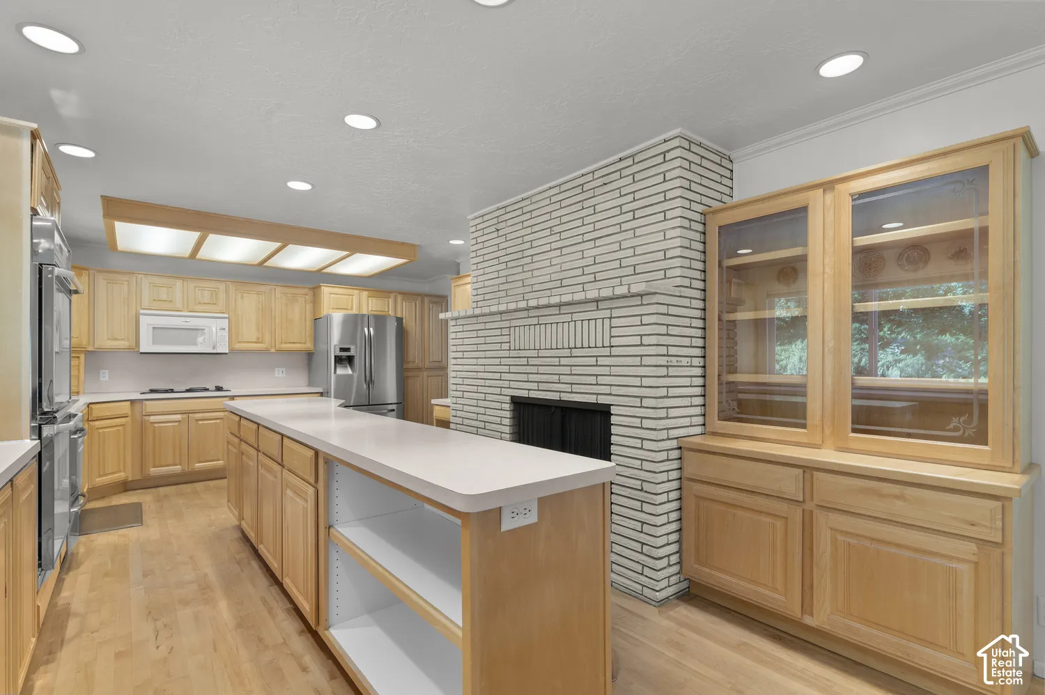 Natural wood cabinetry with integrated storage, white brick fireplace, center island with open shelving, stainless steel appliances, and recessed lighting