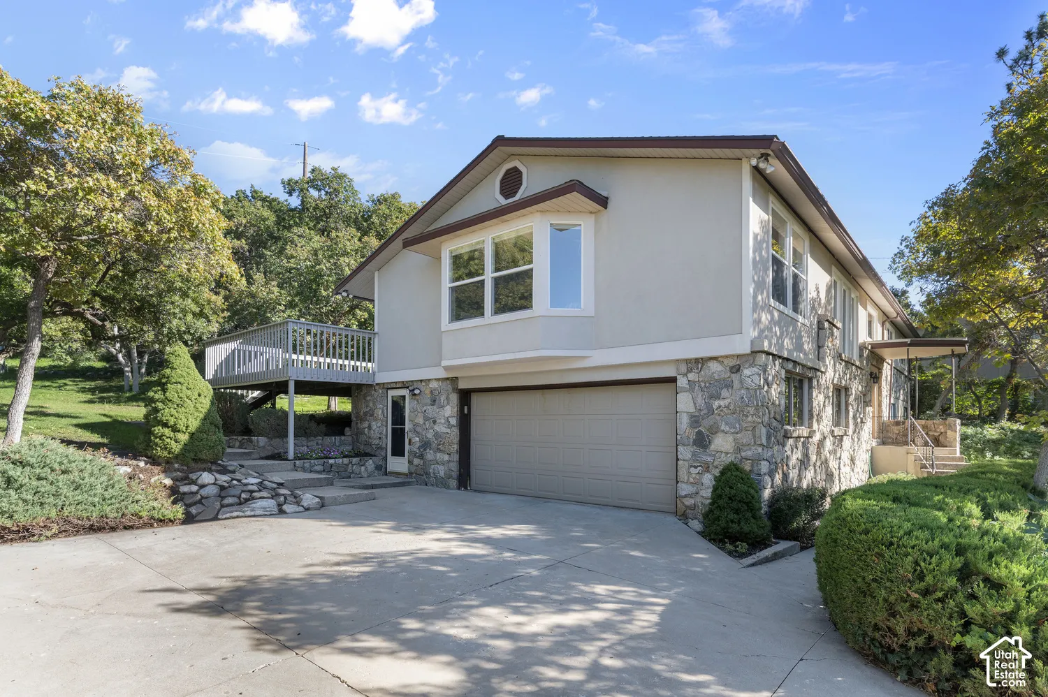 Stone and stucco exterior with an attached garage, upper-level deck, concrete driveway, and mature landscaping
