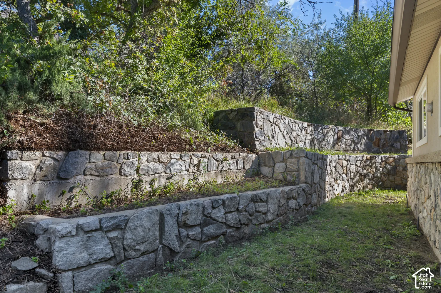 Terraced stone retaining walls with integrated planting beds, bordering a grassy area beside the property