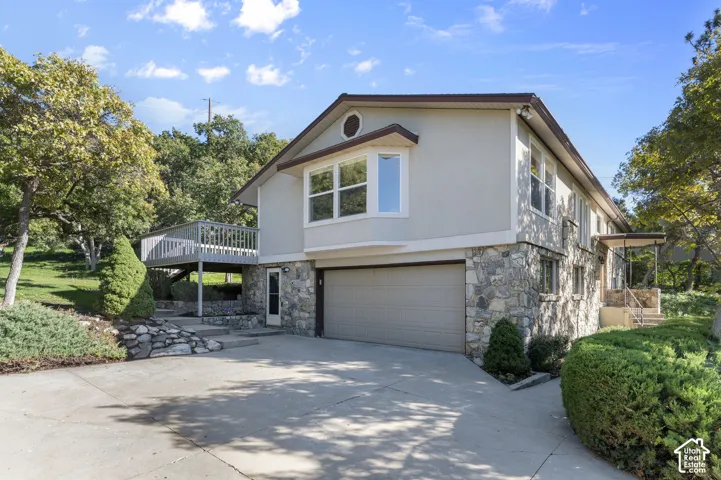 Stone and stucco exterior with an attached garage, upper-level deck, concrete driveway, and mature landscaping