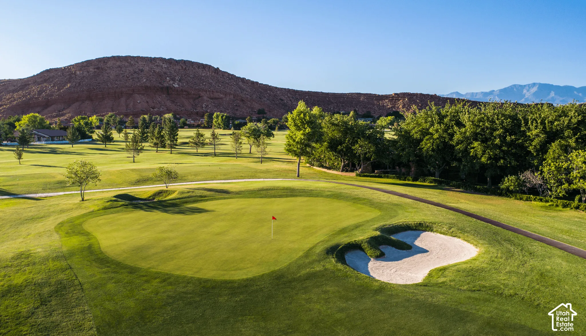 Manicured golf course green with a red flag, surrounded by a sand trap, mature trees, and distant mountainous terrain