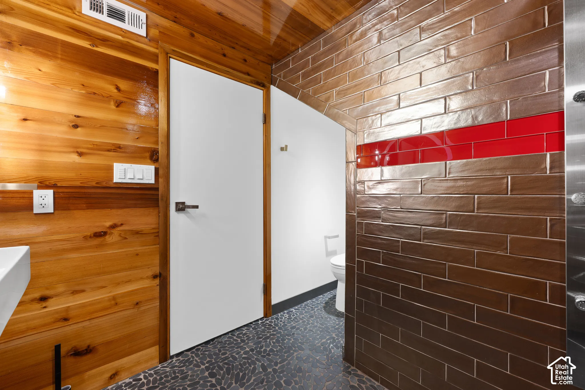 Wood-paneled wall, modern white vanity, textured floor tiling, contrasting wall tile with red accent, and contemporary door hardware