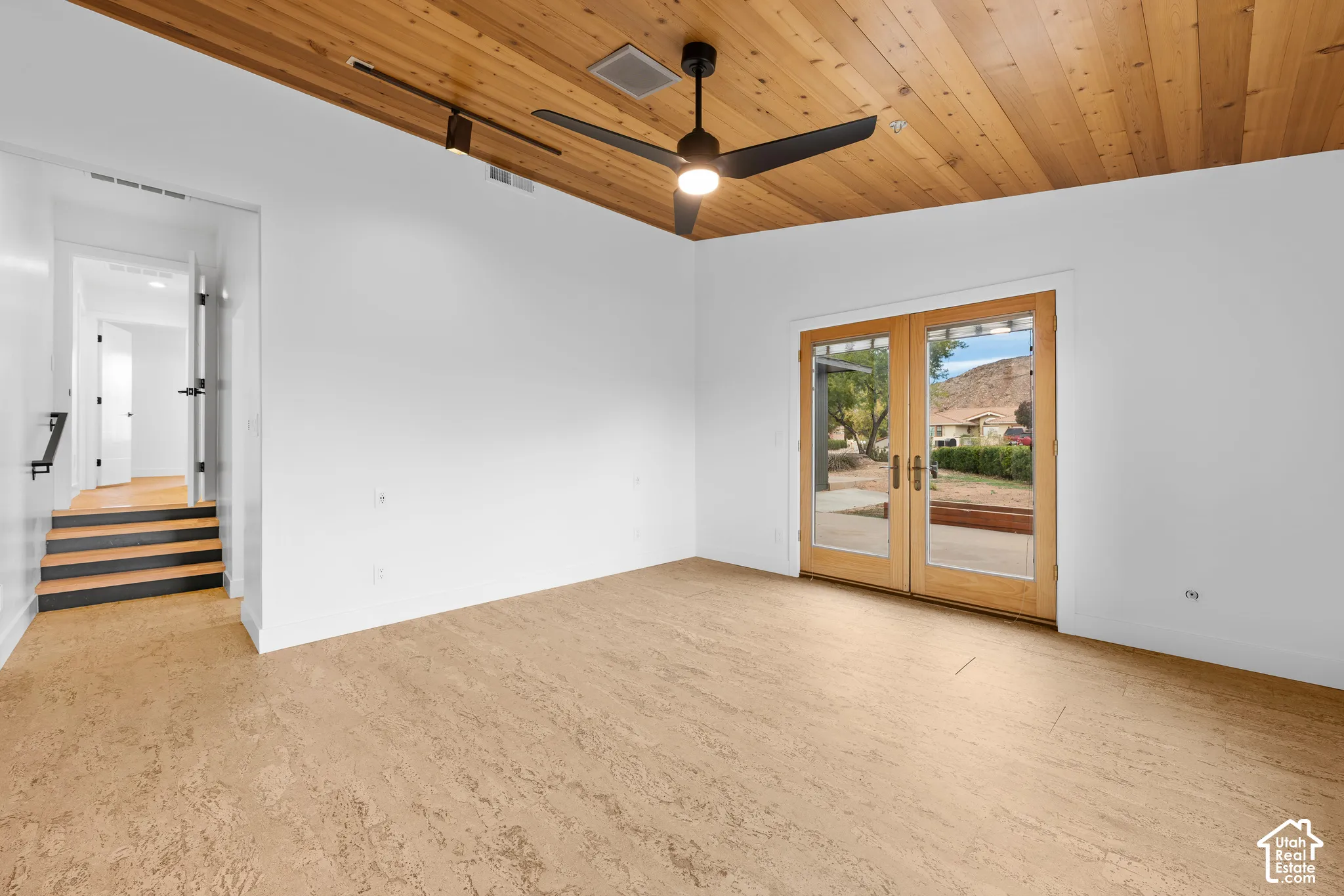 Wood-paneled ceiling with a ceiling fan, light-toned flooring, light wood French doors, and a partial staircase with wood treads and a black handrail