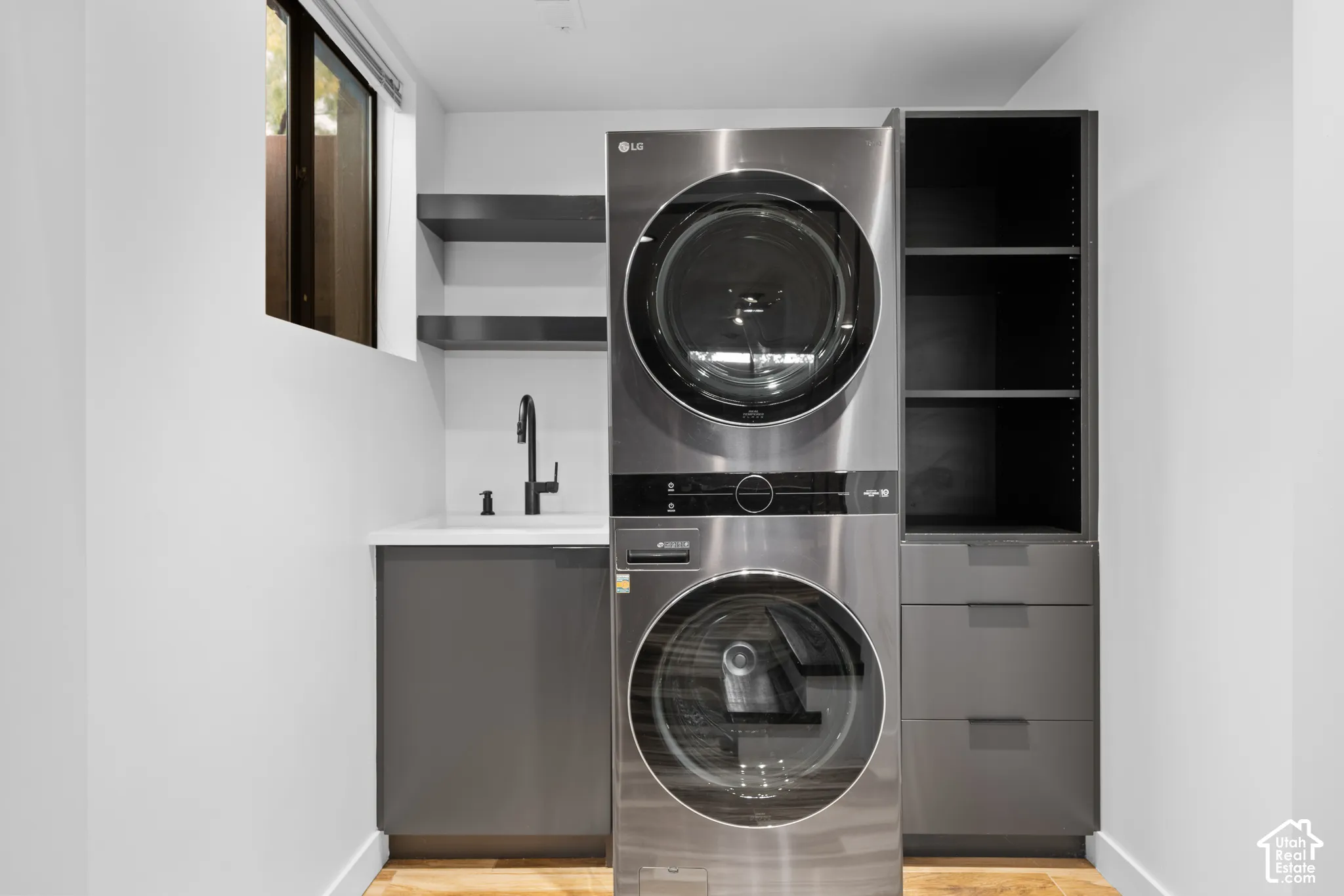 Stacked stainless steel washer and dryer, built-in shelving, under-sink cabinetry, and a modern wall-mounted faucet