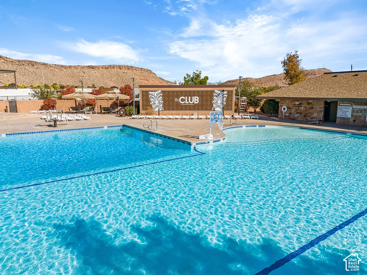 Expansive outdoor pool with integrated shallow area, surrounded by a paved deck, and a clubhouse building featuring stone-look siding and a shingle roof