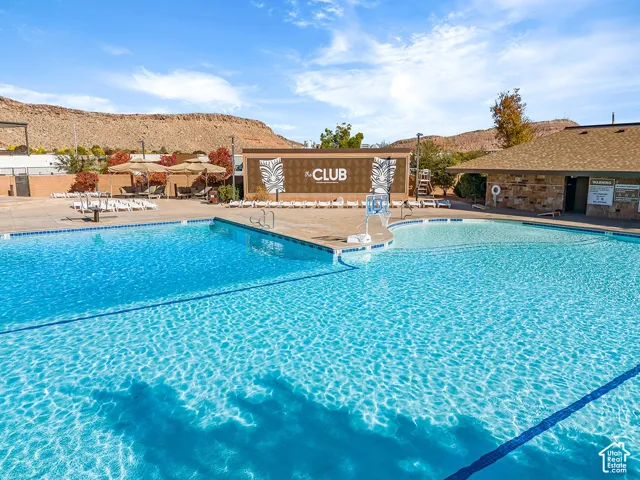 Expansive outdoor pool with integrated shallow area, surrounded by a paved deck, and a clubhouse building featuring stone-look siding and a shingle roof