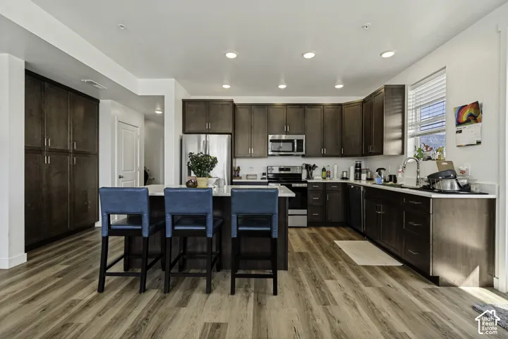 Kitchen featuring wood-finish flooring, stainless steel appliances, dark wood cabinetry, white countertops, and a central island with bar seating