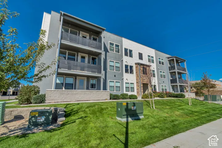 Modern multi-story building featuring balconies with horizontal railings, mixed-material facade with stone accents, and a sloped, manicured lawn