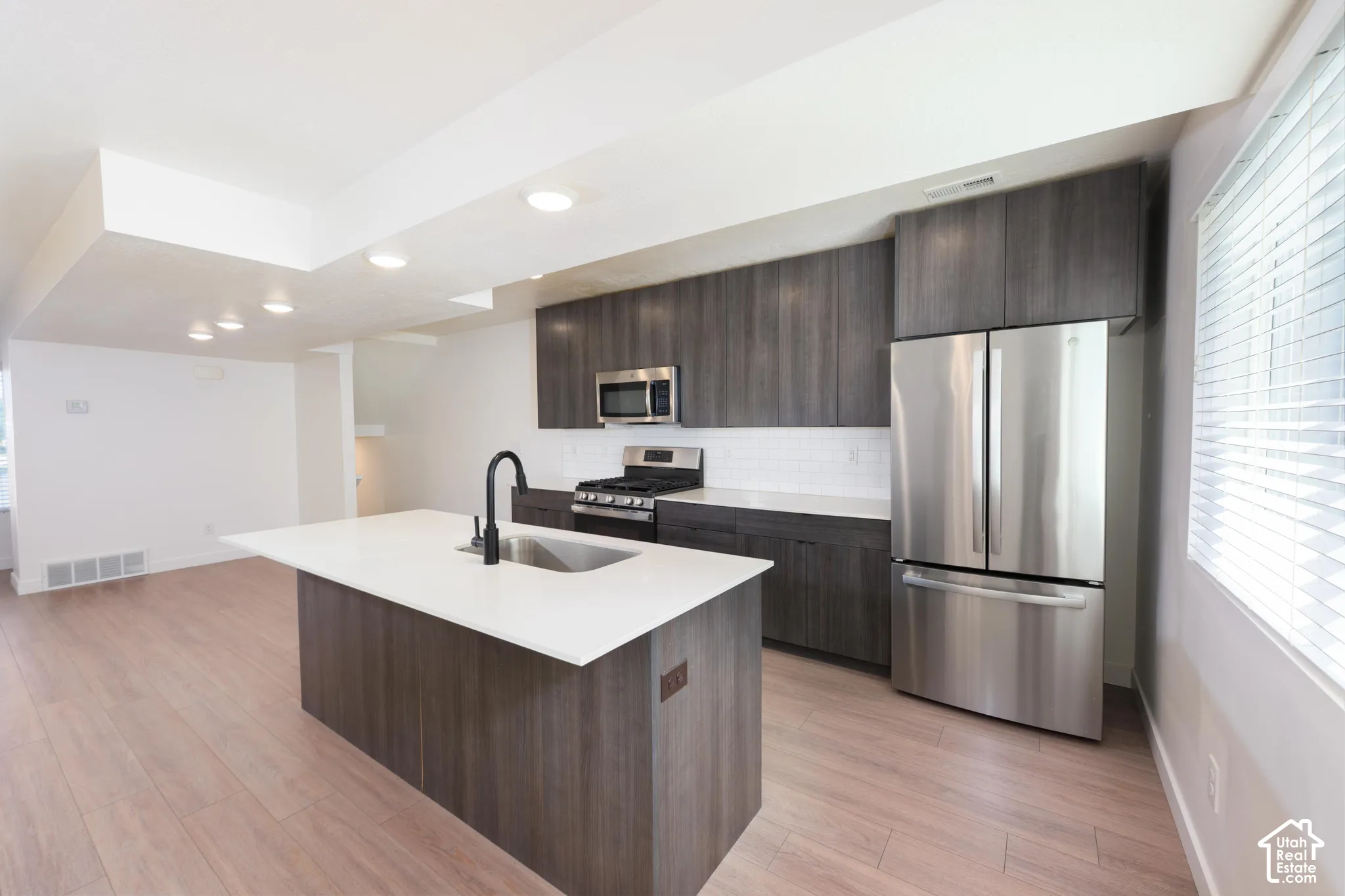 Kitchen featuring dark wood-finish cabinetry, stainless steel appliances, a white subway tile backsplash, and a large island with a composite countertop and integrated sink