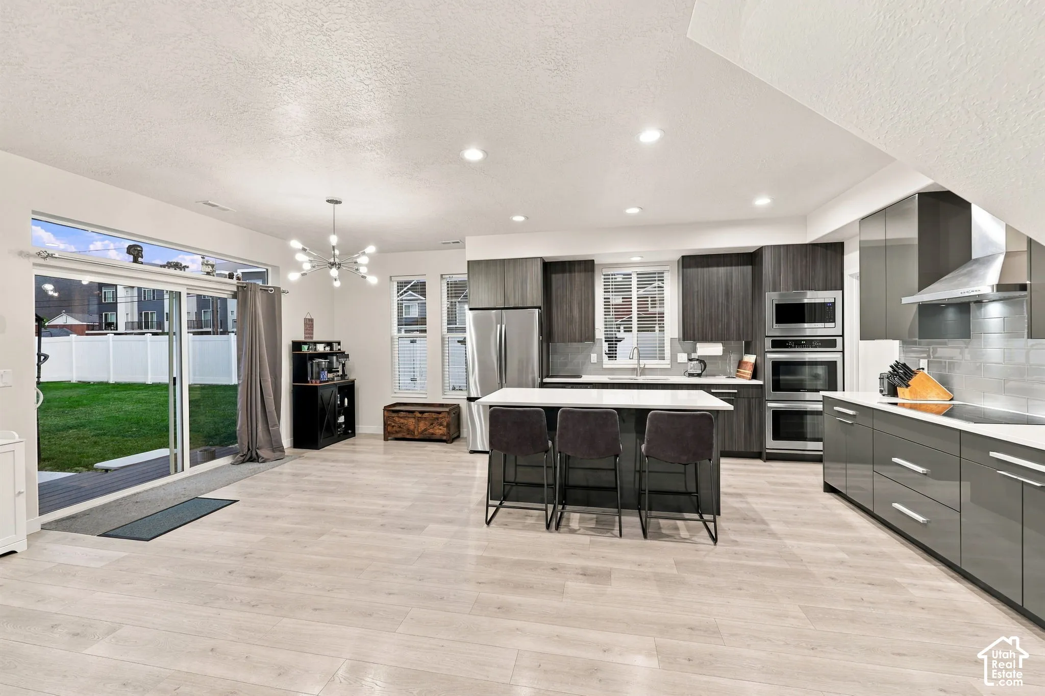 Open-concept living space with light wood-finish flooring, modern kitchen, white countertops, built-in stainless steel appliances, and a sliding glass door to the backyard