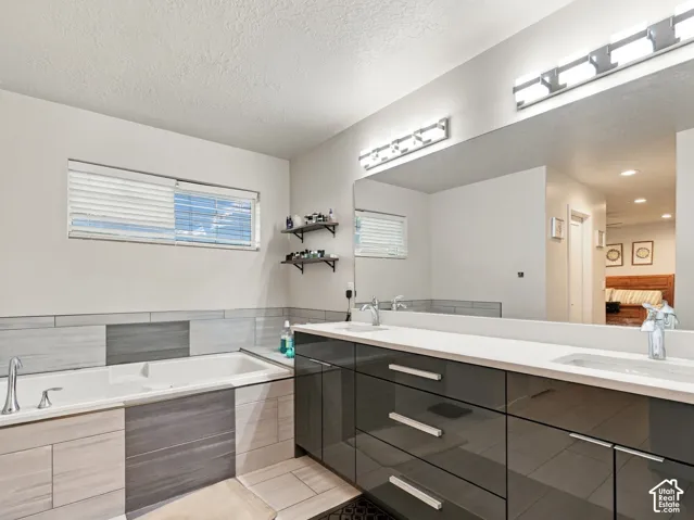 Built-in soaking tub with surrounding tile, dual vanity with undermount sinks, expansive mirror, wall-mounted lighting, and a high-gloss vanity with drawer and cabinet storage