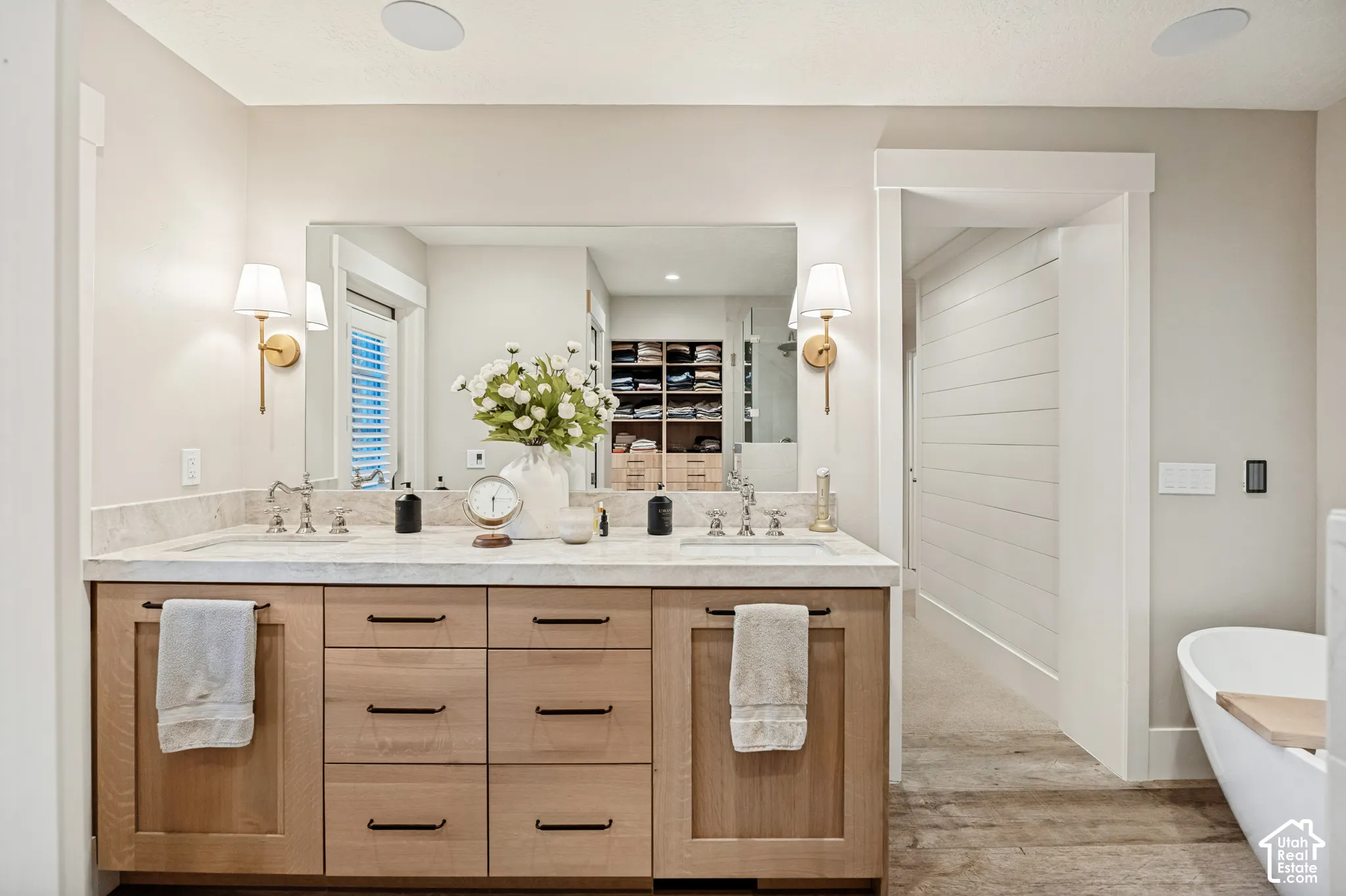 Dual vanity with light wood-finish cabinetry, stone countertop, and brushed brass sconces