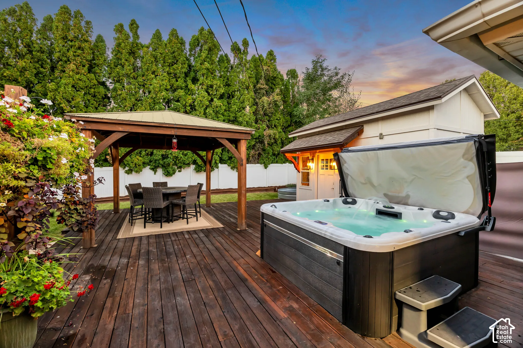 Expansive wood-finish deck featuring a built-in spa, a separate wood gazebo with a metal roof, and a detached outbuilding with exterior lighting