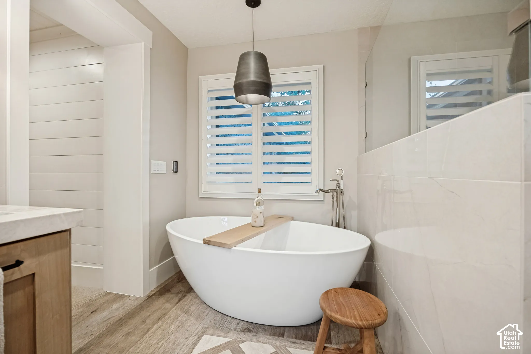 Freestanding soaking tub with a modern pendant light, heated wood/tile flooring, and white plantation shutters
