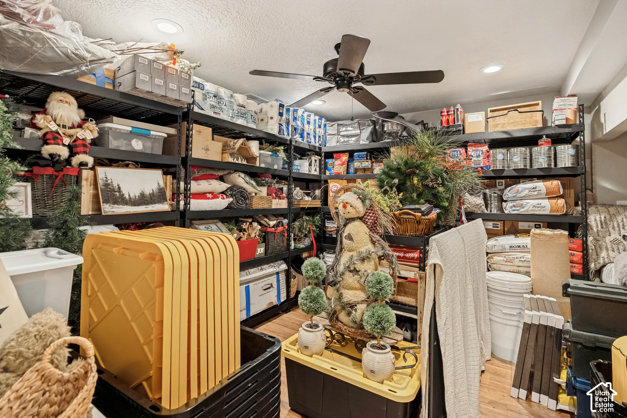 Recessed ceiling lights, wood-finish flooring, and a ceiling fan.  Used as a storage room but is a bedroom with window behind the storage shelves.