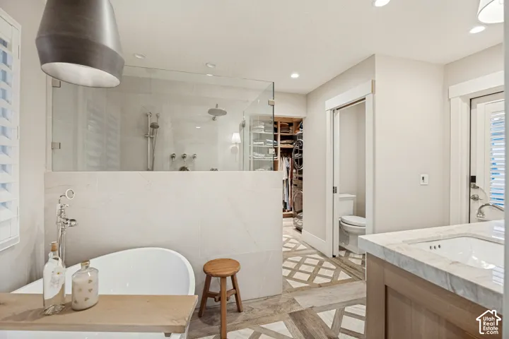 Freestanding soaking tub, walk-in shower with clear glass enclosure, custom built-in closet system, single vanity with integrated sink and stone countertop, and decorative heated wood tile flooring