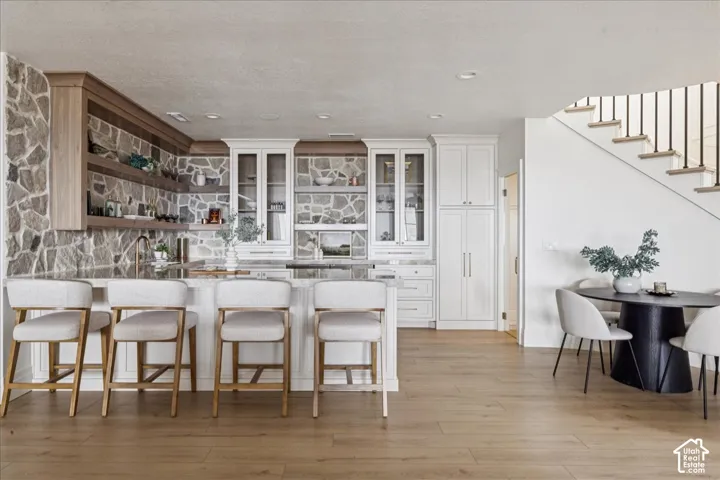 Light wood-finish flooring, stone accent wall, white cabinetry with glass-front uppers, kitchen island with seating, and open-tread staircase with metal balusters