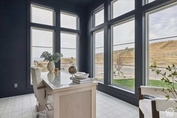 Full-height windows with dark trim, dark wall paint, light-toned flooring, and a light wood-finish desk with metal hardware
