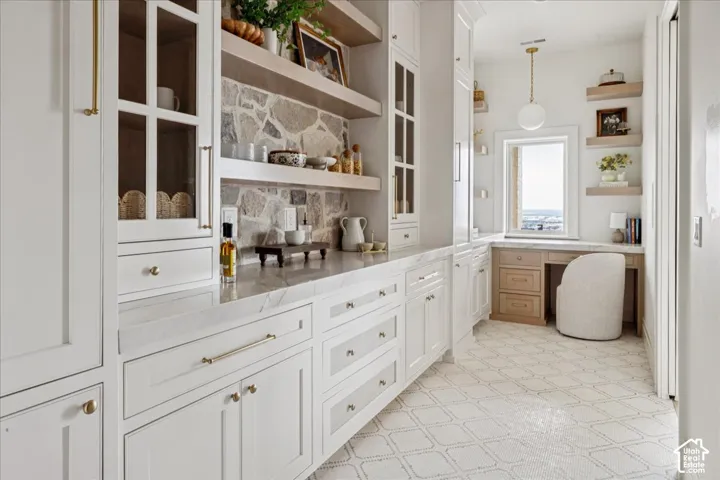 White cabinetry with gold-tone hardware, stone backsplash, built-in desk with wood-finish drawers, pendant lighting, and patterned floor tile