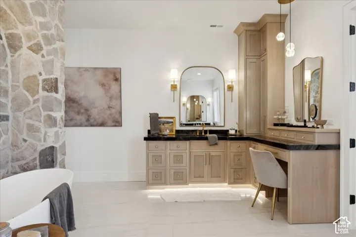 Freestanding tub with a natural stone accent wall, dual vanity with dark countertops, built-in makeup vanity, and recessed lighting